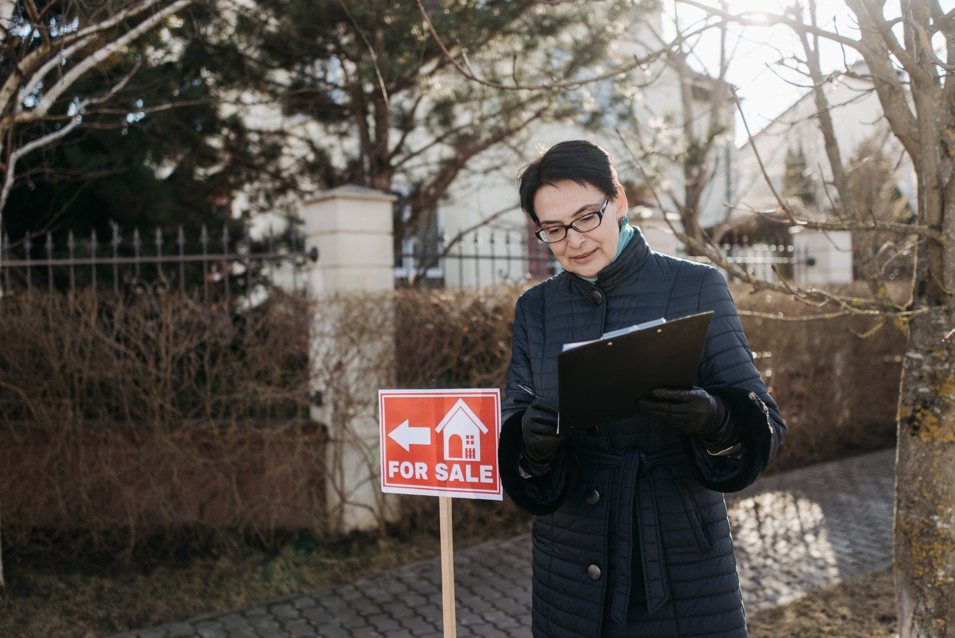 A woman is standing in front of a for sale sign holding a clipboard.