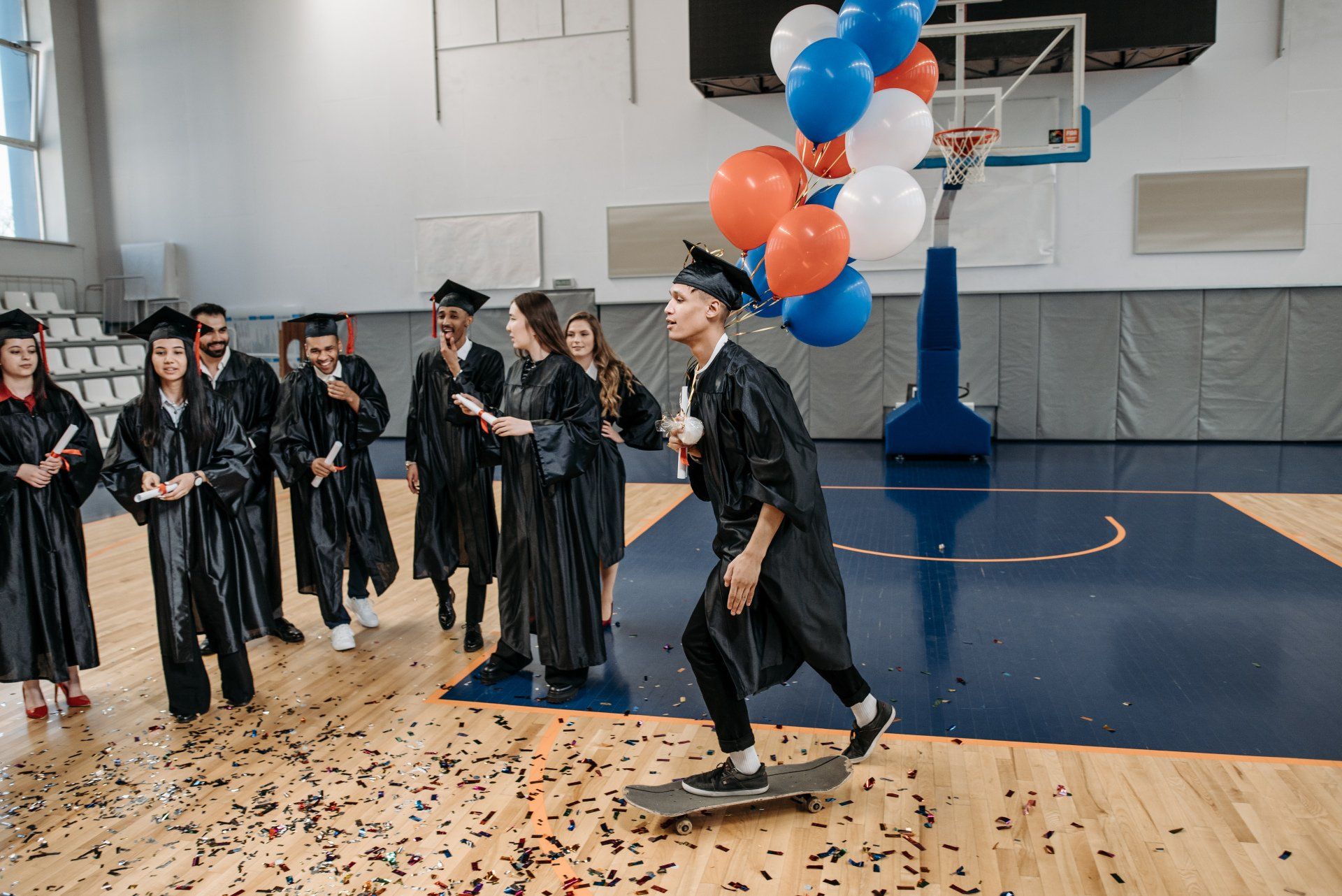 Graduation: Student on skateboard, holding balloons, surrounded by graduates in a gym.