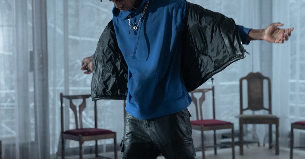 A man in a blue hoodie and black jacket is dancing in a room with chairs.