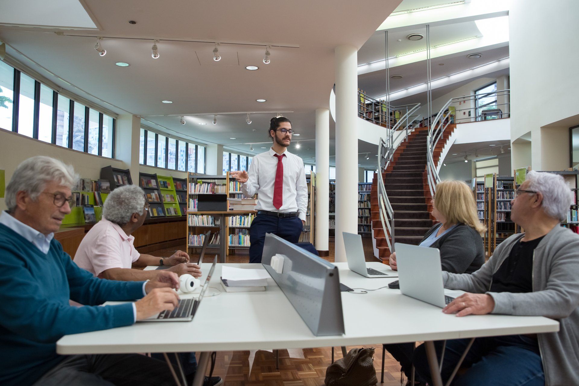 A man leads a computer class for seniors in a library.  Students use laptops at a large table.