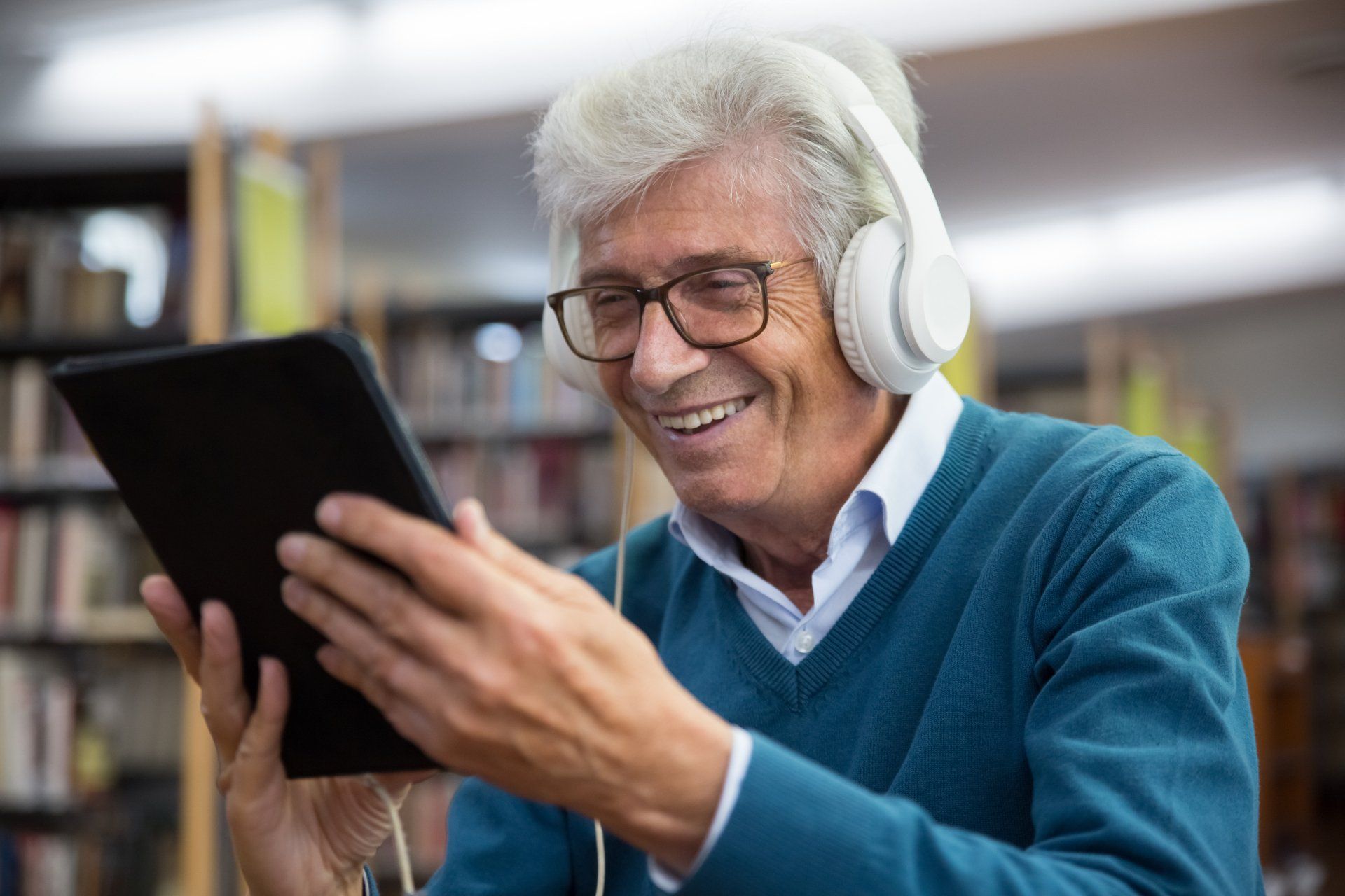 An elderly man wearing headphones is using a tablet computer in a library.