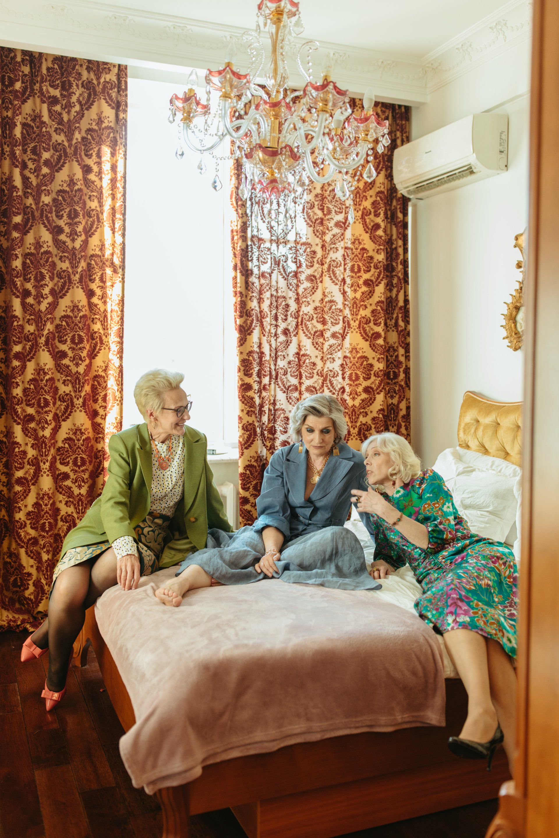 Three older women are sitting on a bed in a bedroom.