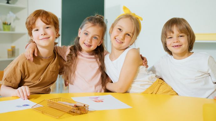 A group of children are sitting at a table with their arms around each other.