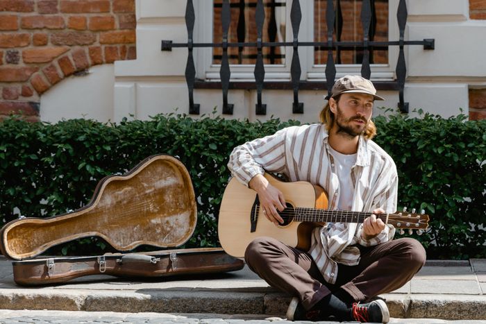 A white man sitting on a curb, strumming a guitar with the case opened next to him