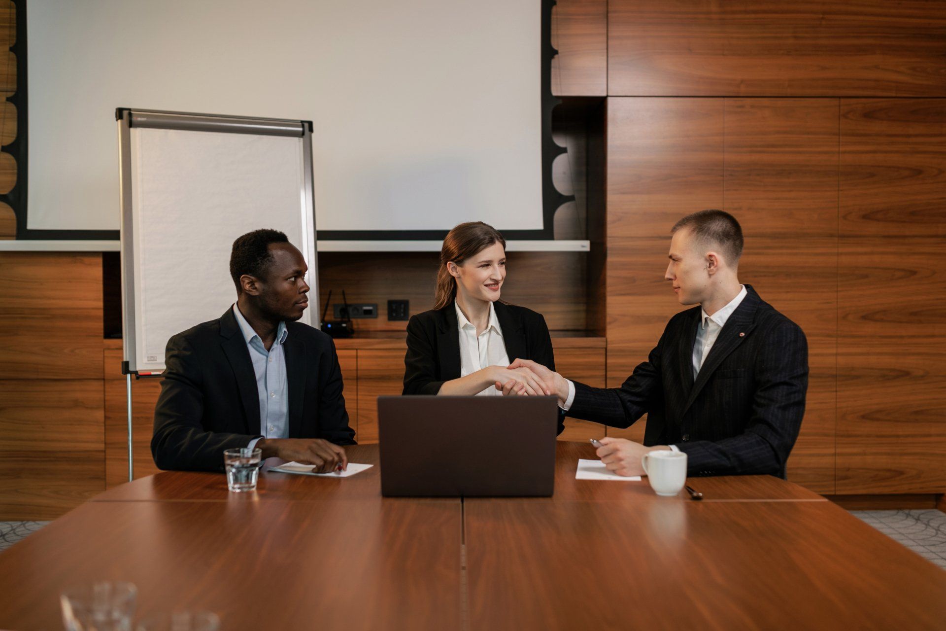 Three people gathered around a laptop in a meeting setting. Two people are shaking hands.