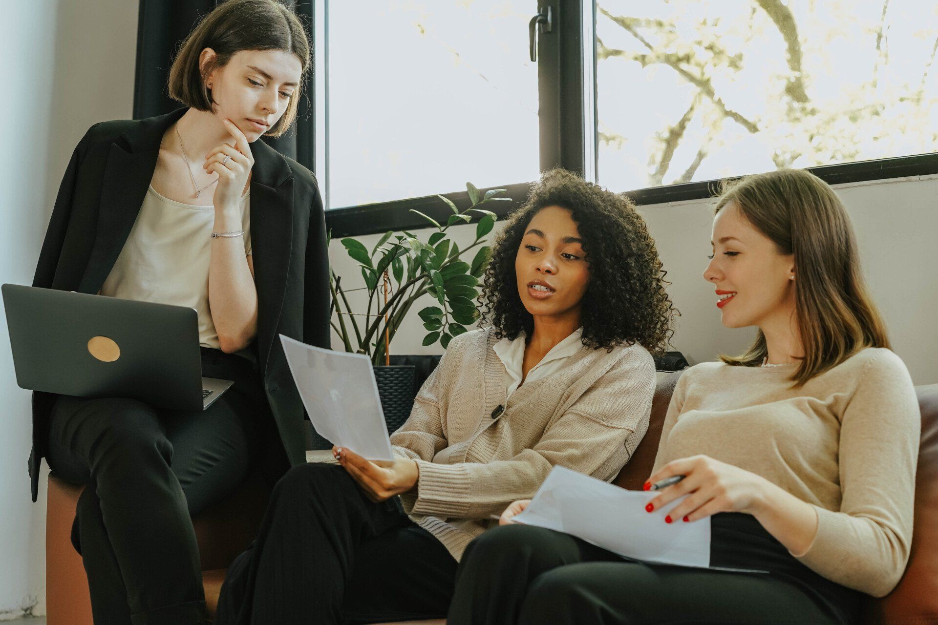 Three women are sitting on a couch with laptops and papers.