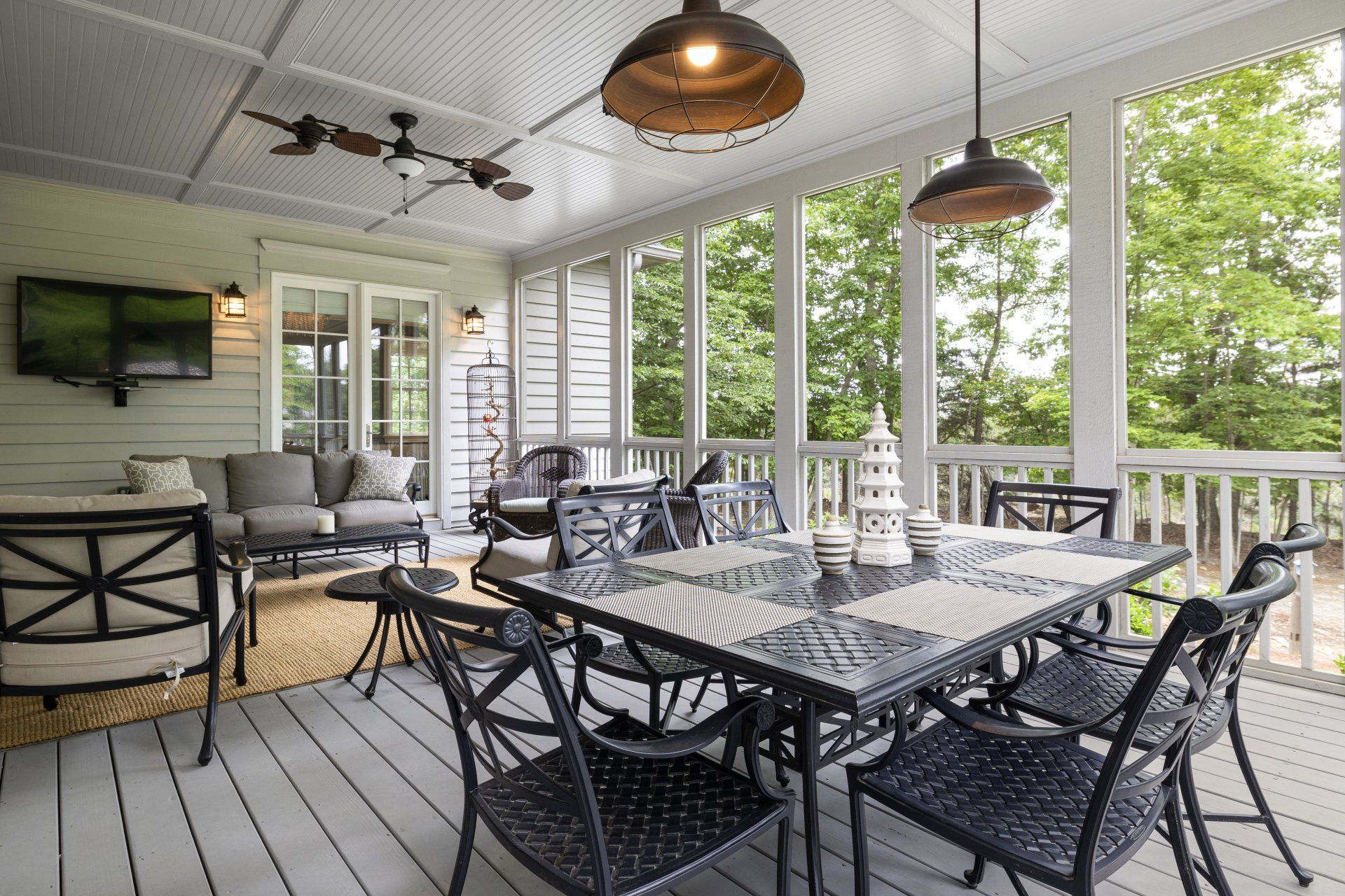 Screened-in porch with dining table and seating. Green trees visible through windows, light fixtures hang from the ceiling.