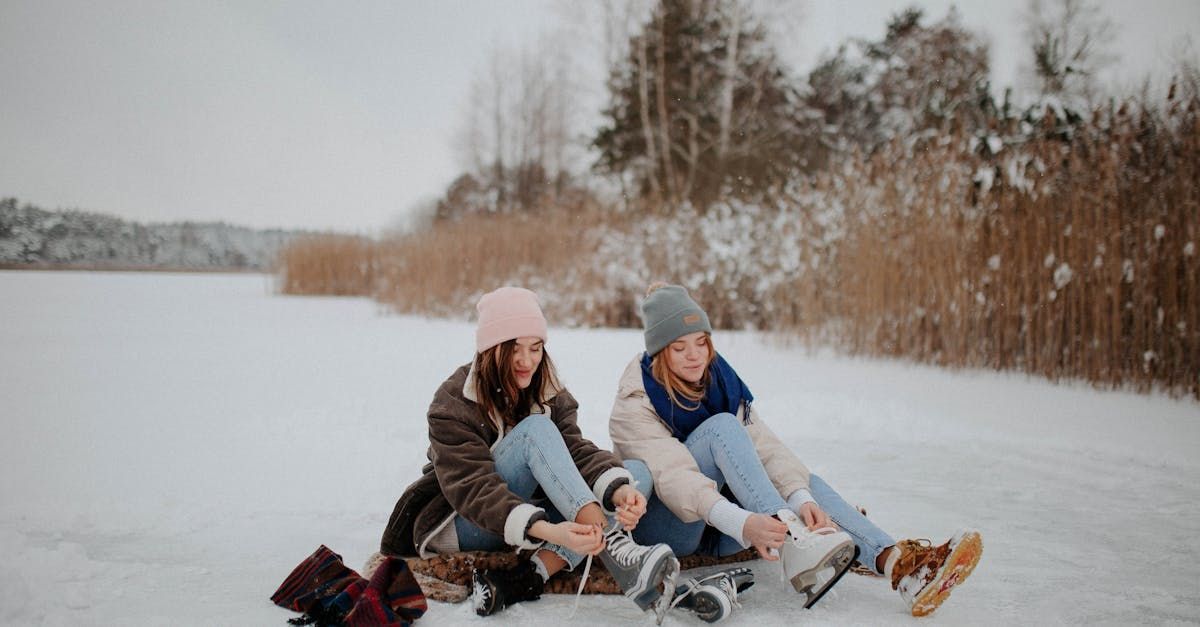 Two women are sitting in the snow tying their ice skates.