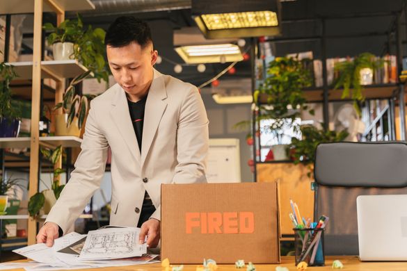 A person in a beige blazer packs office papers into a cardboard box labeled "FIRED" on a desk in a modern office space.