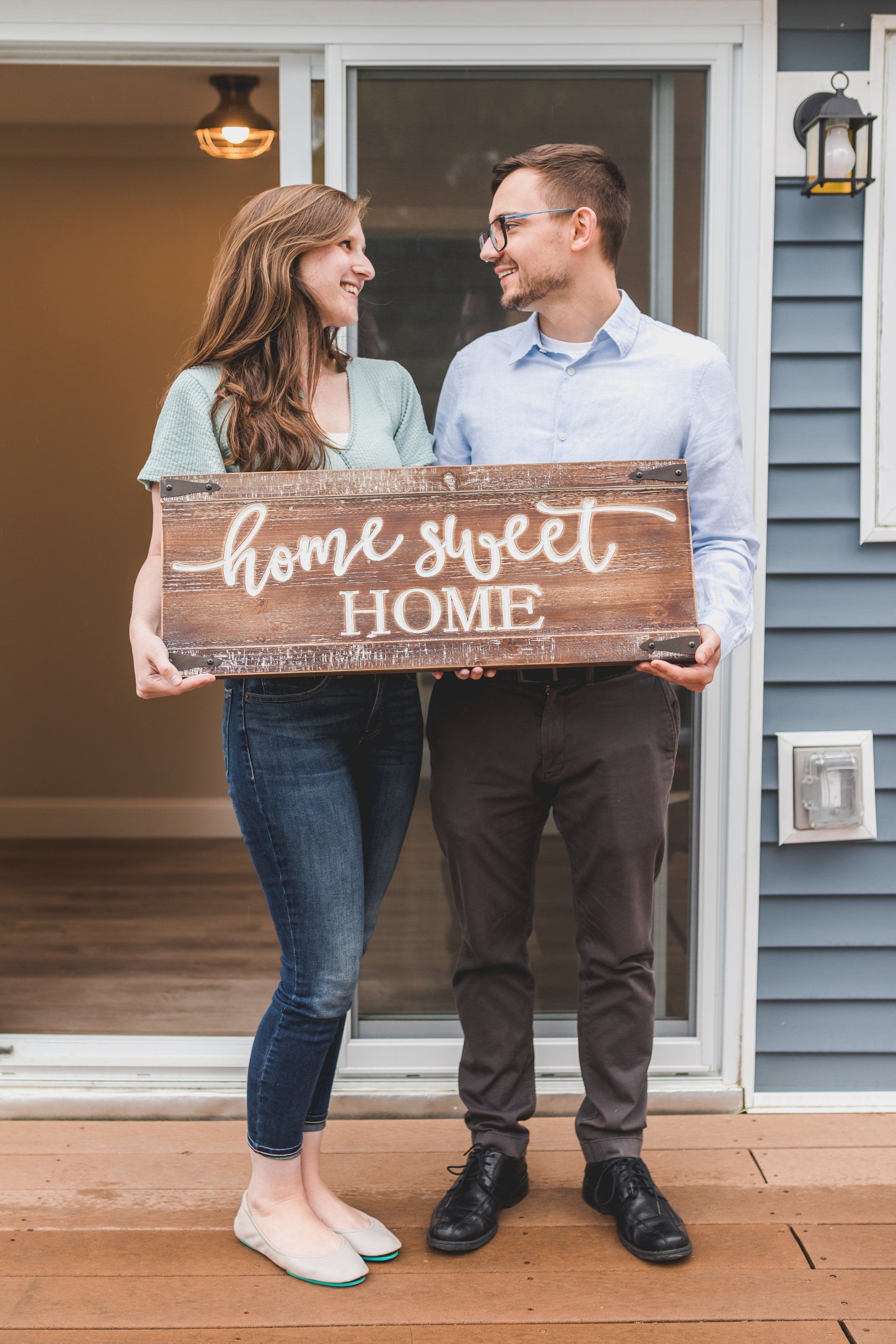 A happy couple stands in front of a house, holding a wooden sign between them.