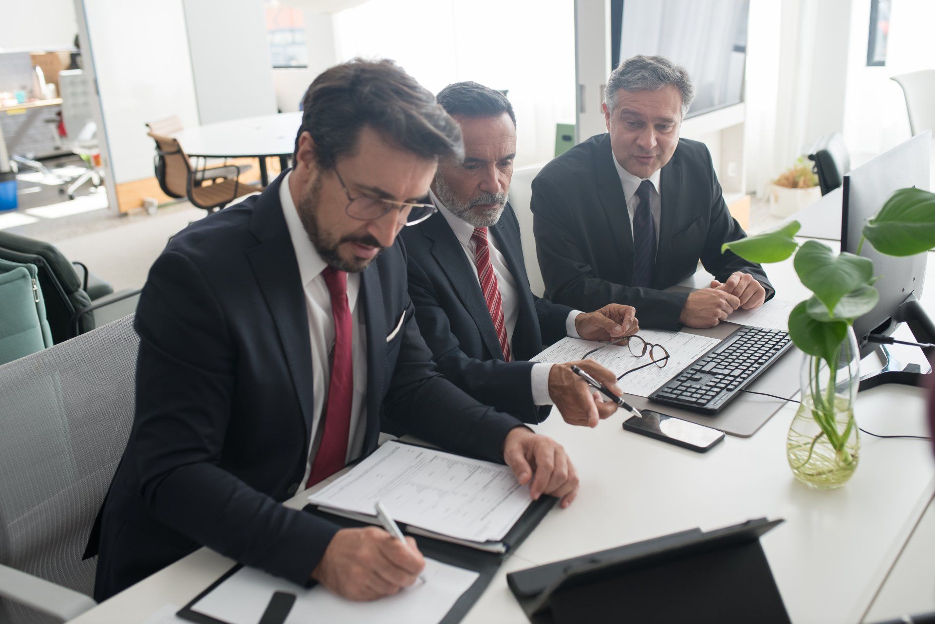 três homens de terno e gravata estão sentados em uma mesa em frente a um computador.