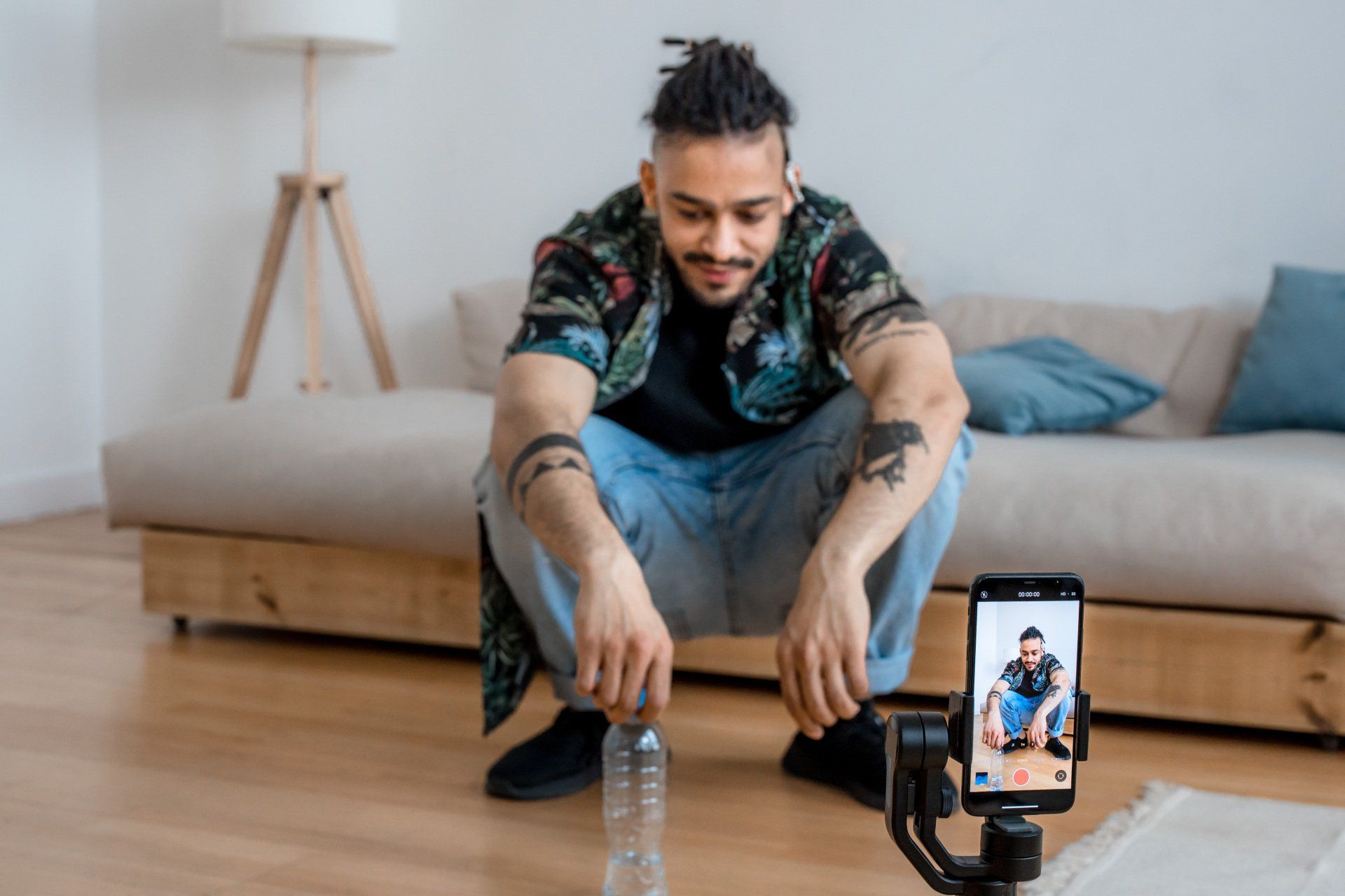 A man is squatting down in front of a camera while holding a bottle of water.