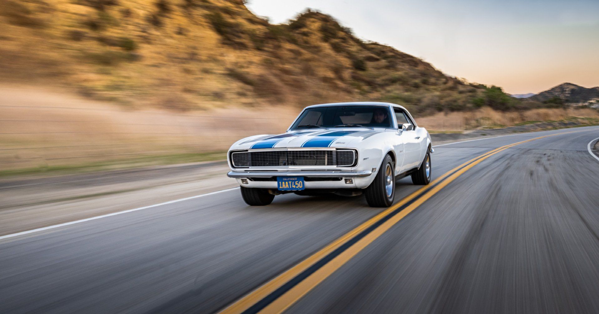 A white and blue muscle car is driving down a road.