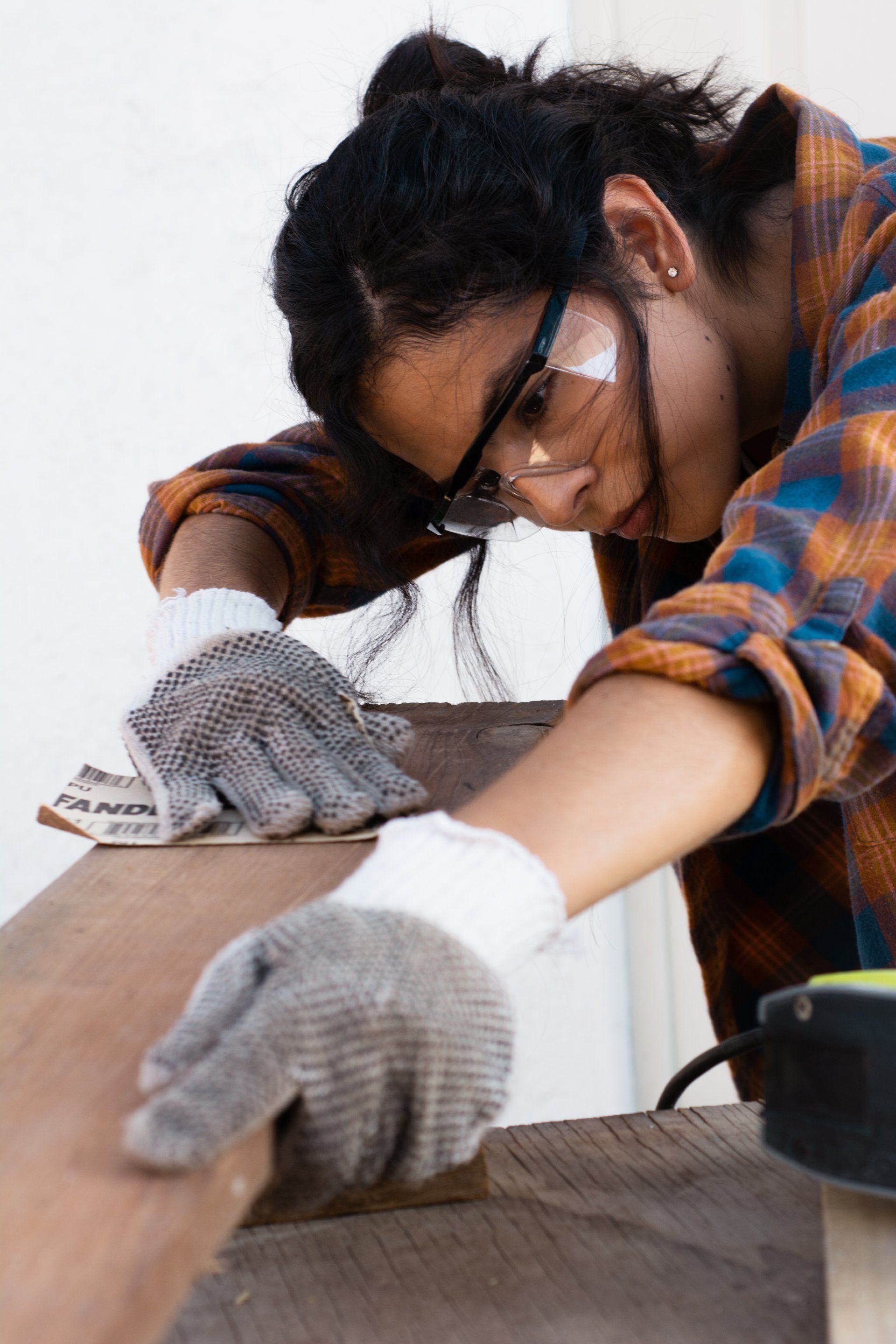 A woman wearing safety glasses and gloves is sanding a piece of wood.
