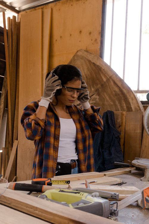 A woman wearing headphones and goggles is standing in a woodworking shop.