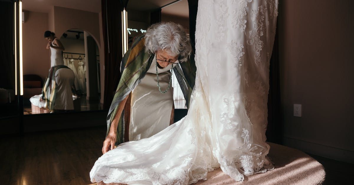 An elderly woman is trying on a wedding dress in front of a mirror.