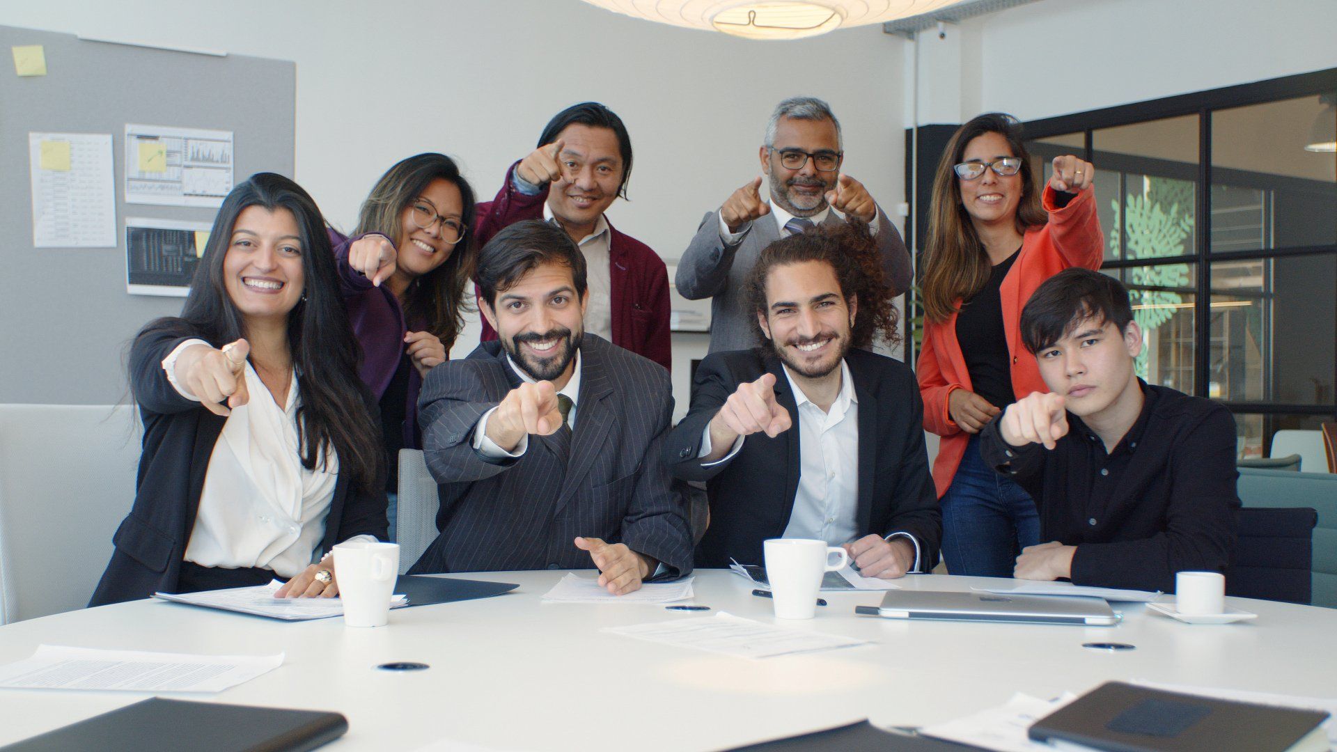 A group of people are sitting around a table and pointing at the camera.