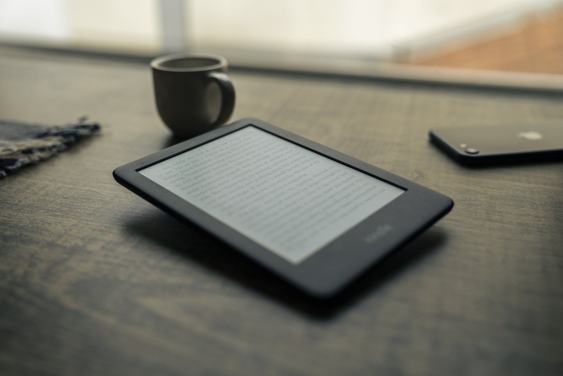 An e-reader lays on a table top with a mug and a smartphone in the background.