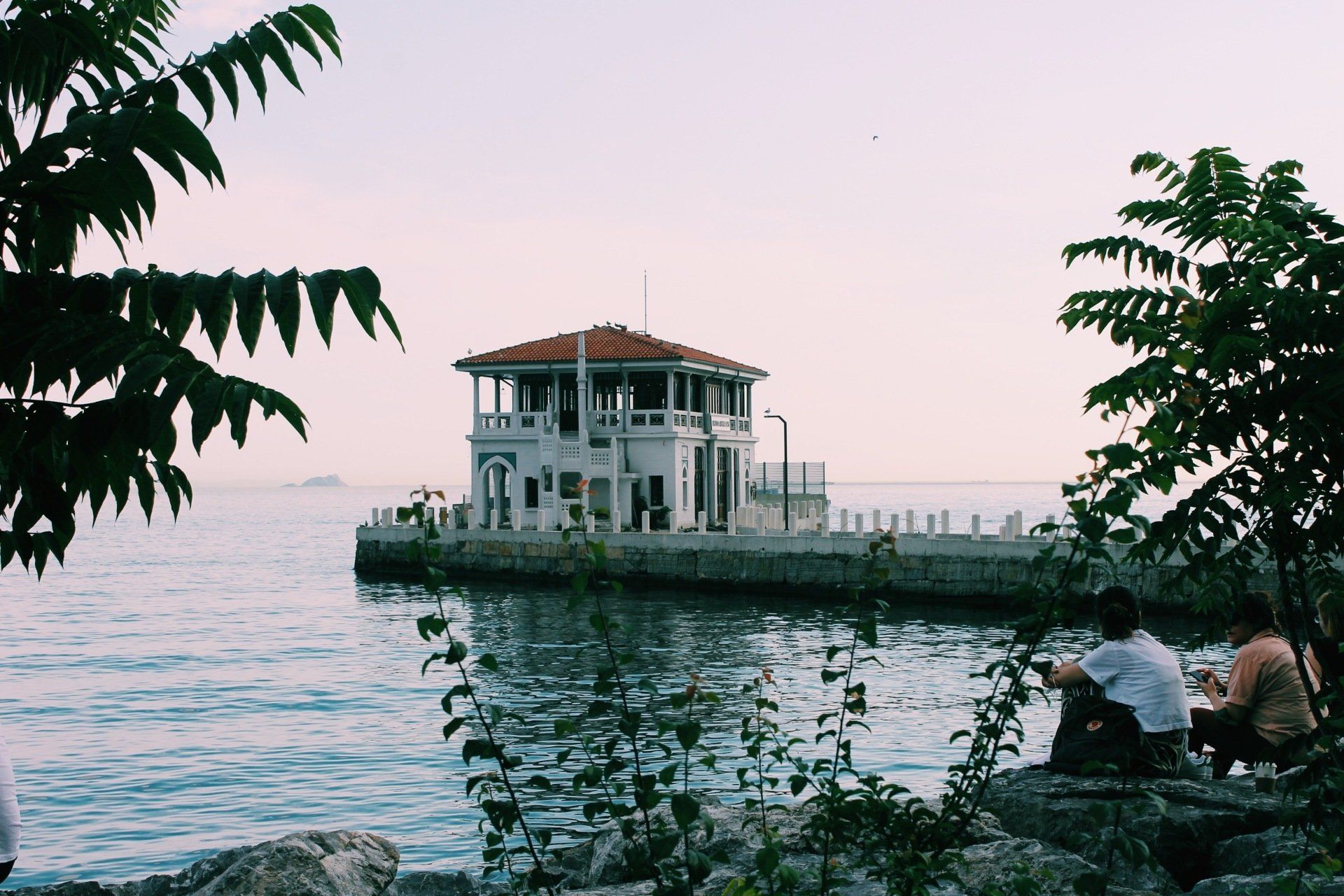 A couple sits on a rock near a body of water with a dock and small house in the background