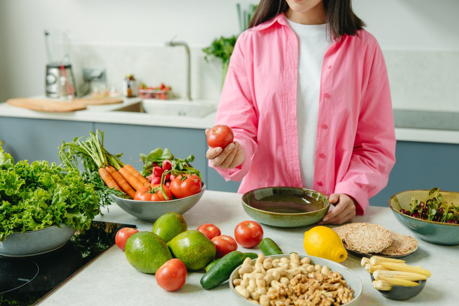 Een vrouw in een roze shirt houdt een tomaat vast voor een tafel vol fruit en groenten.