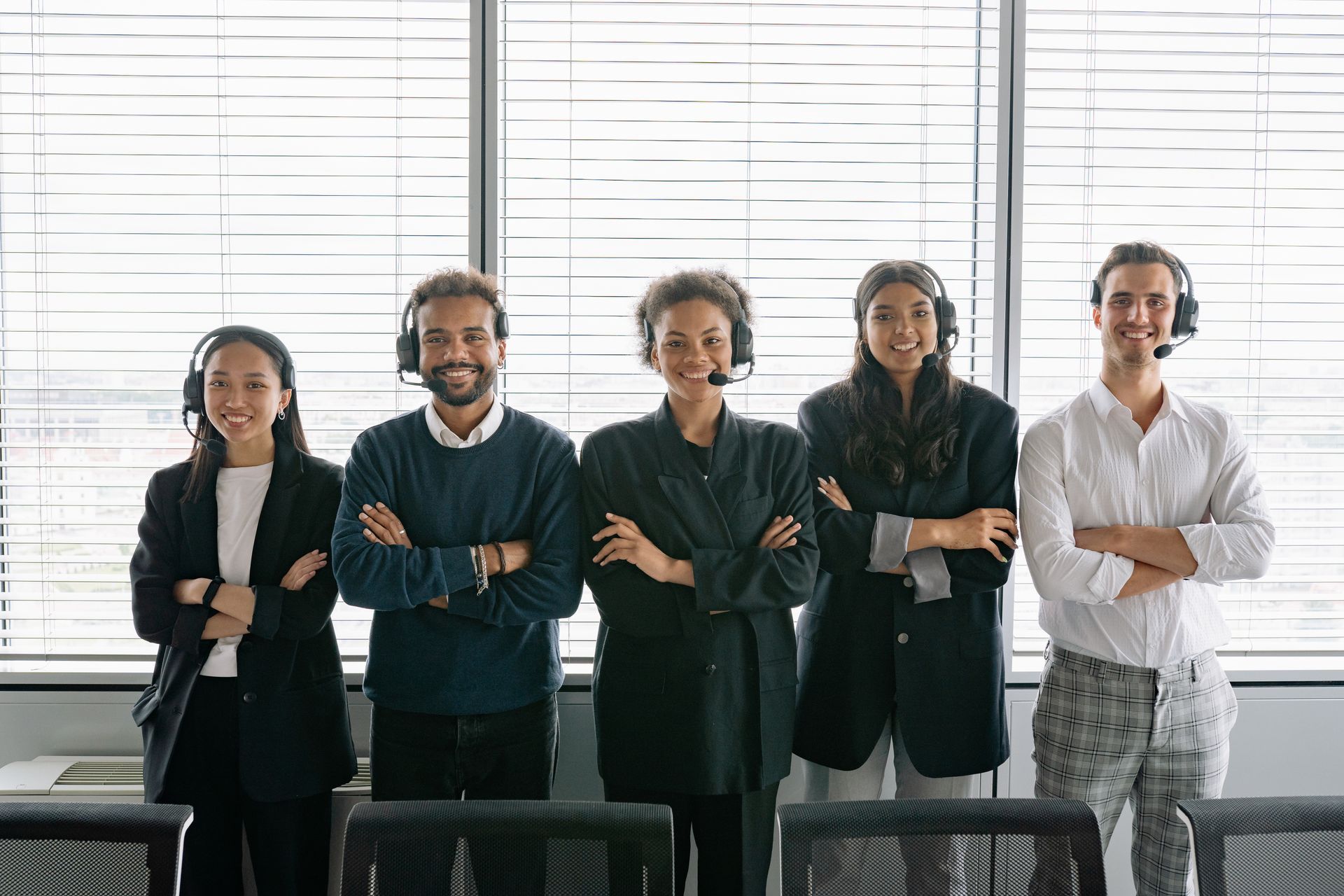 Five people with headsets, arms crossed, smiling, in front of a window with blinds; likely a call center setting.