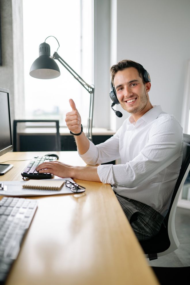 A man wearing a headset is sitting at a desk with a computer and giving a thumbs up.