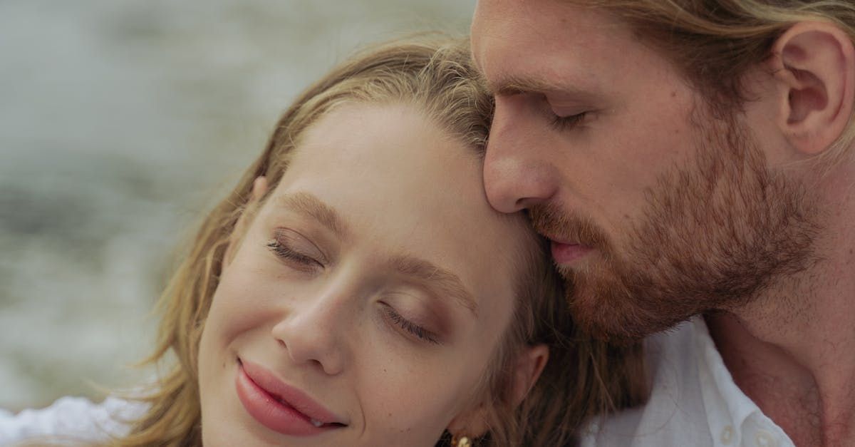 Woman with closed eyes rests head against a man's, both with eyes closed, near water.