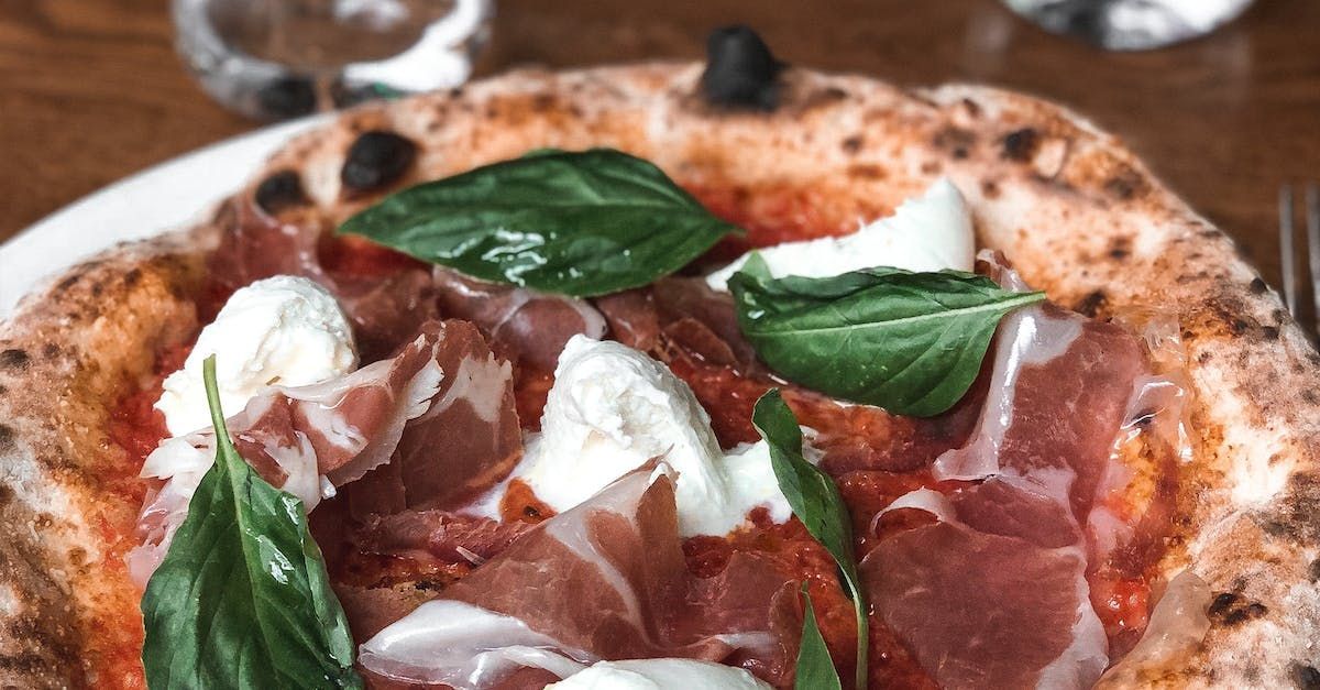 Close-up of a pizza featuring a delicious combination of fresh ingredients. The pizza is topped with thin slices of prosciutto, dollops of creamy mozzarella, and fresh basil leaves. The crust is golden and slightly charred, adding a rustic touch. The vibrant red tomato sauce peeks through the toppings, making the pizza look both appetizing and flavorful. The image captures the artisanal quality of the pizza, highlighting its fresh and high-quality ingredients.
