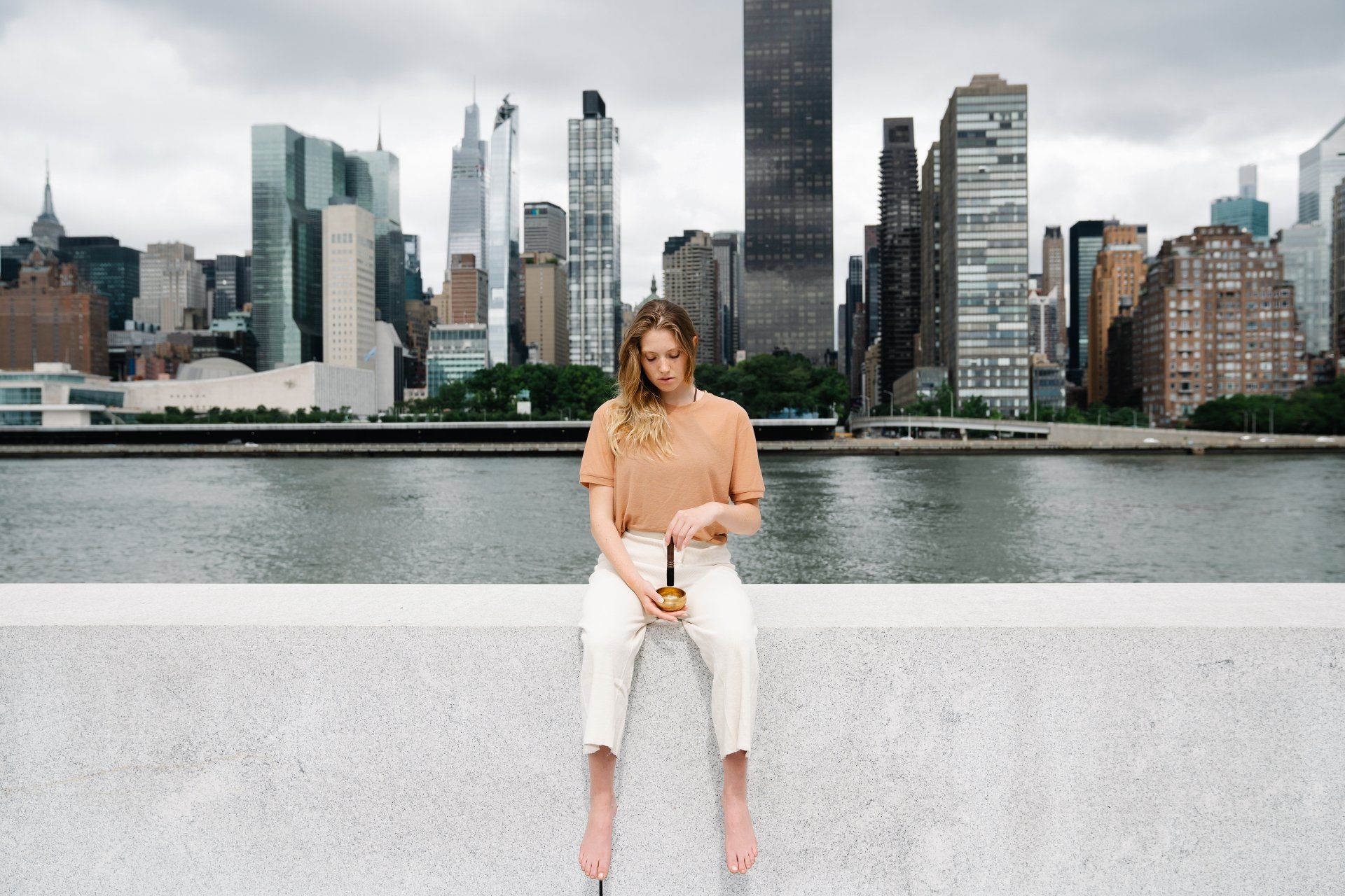 A woman is sitting on a ledge overlooking a body of water with a city skyline in the background.