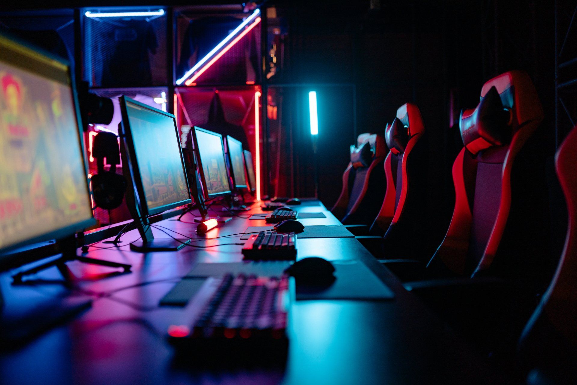 A row of racing-style gaming chairs at a long black desk in a dark room with red and purple led lights. On the desk is one PC set up for each gaming chair, with a keyboard and mouse