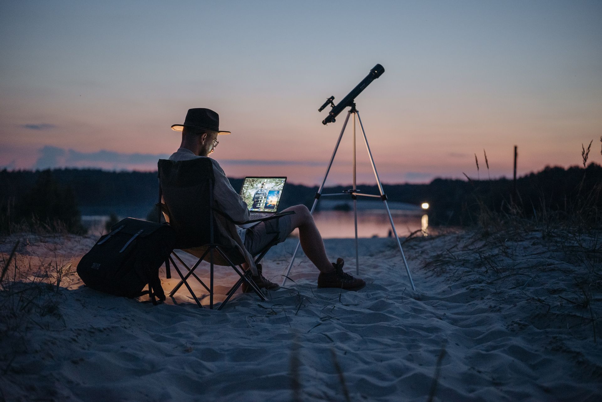 a man is sitting on a beach using a laptop and a telescope .