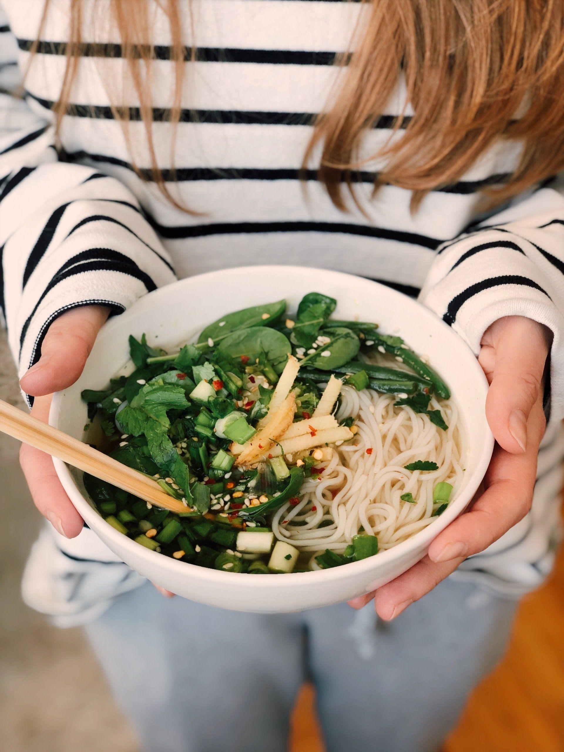 Woman holding a white bowl of Asian noodles with green vegetables, chopsticks.