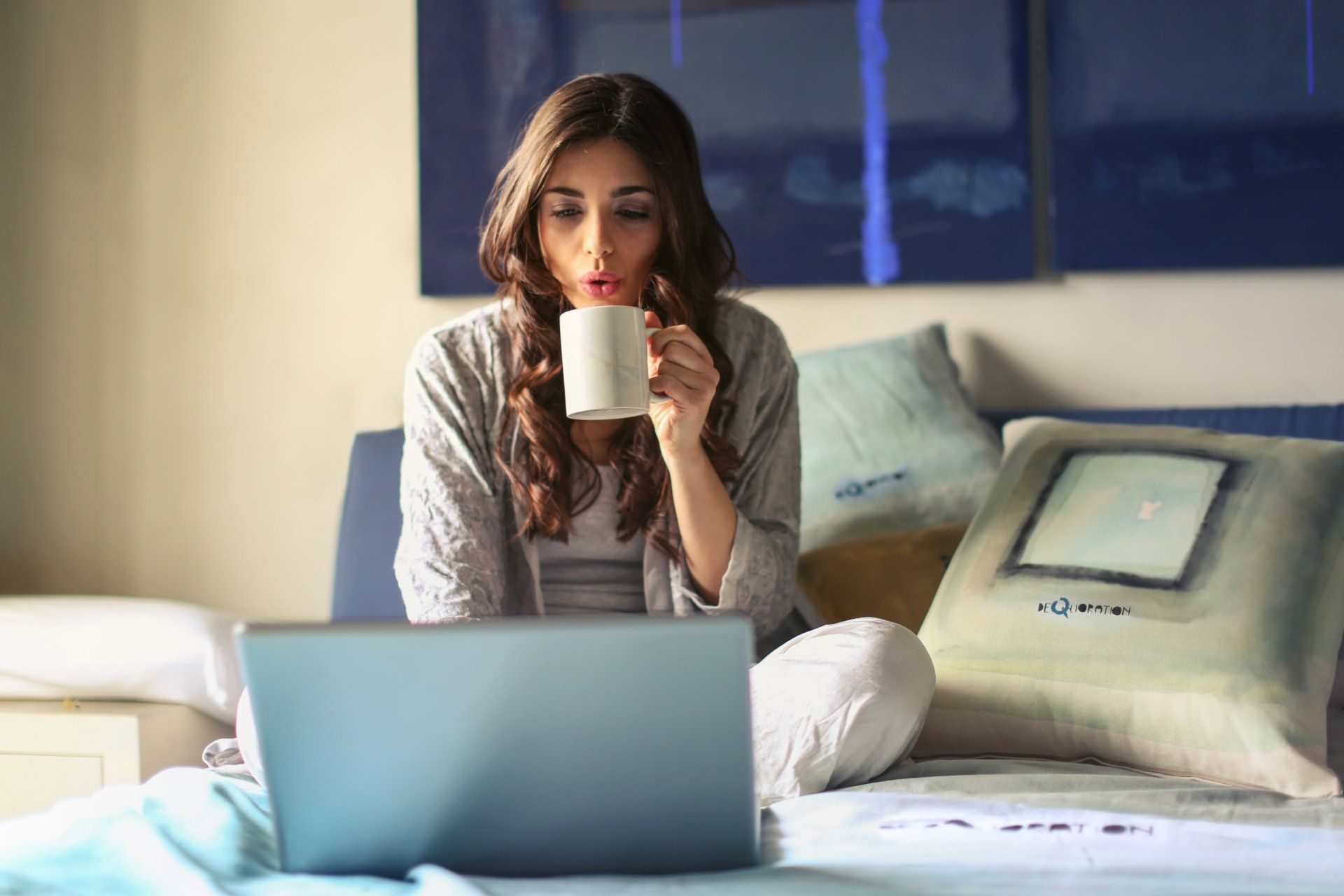 a woman is sitting on a bed drinking coffee and using a laptop computer .