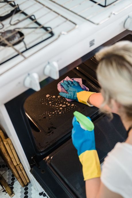 A woman is cleaning an oven with a cloth and spray bottle.