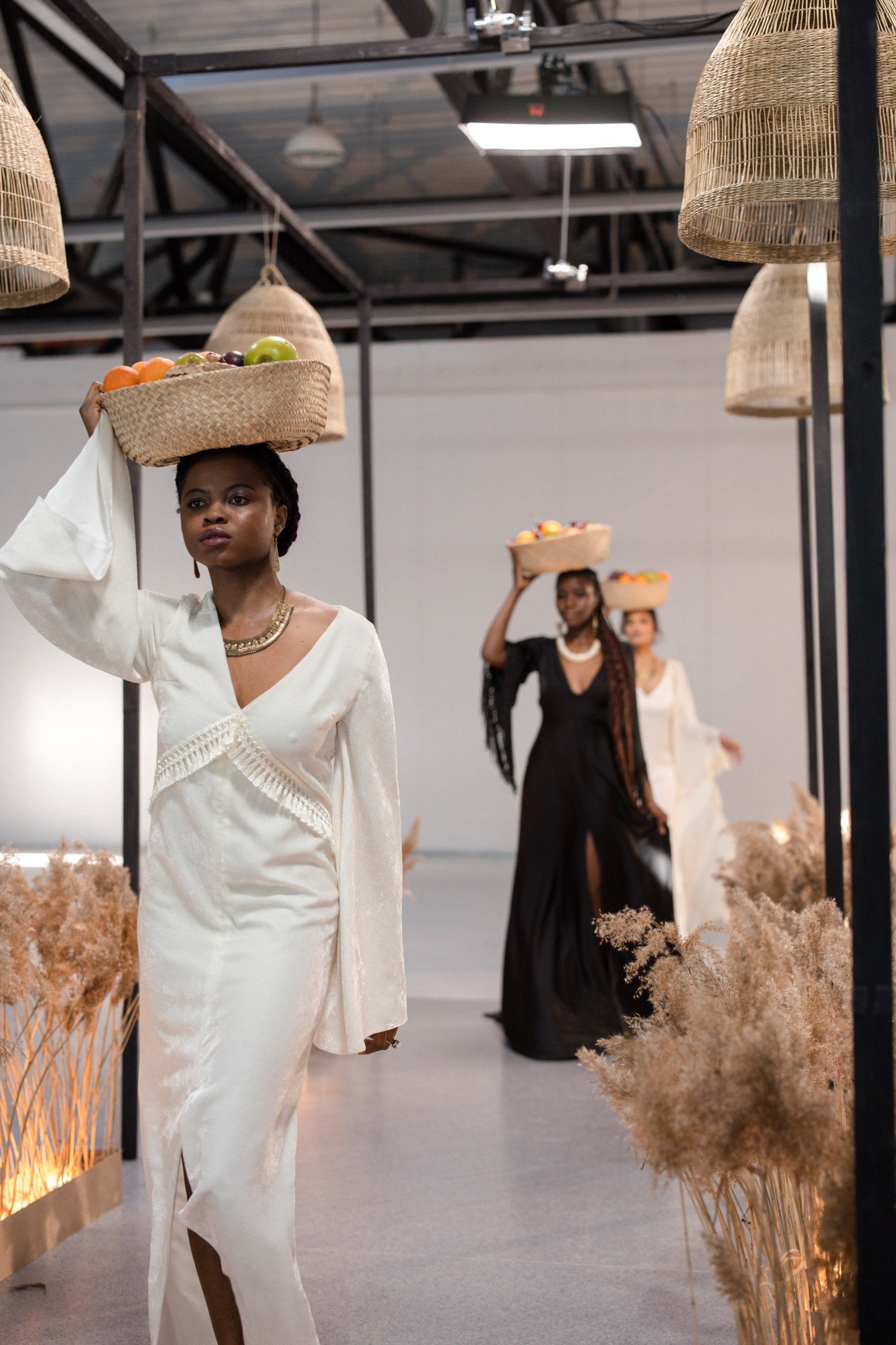 A woman in a white dress is carrying a basket of fruit on her head.
