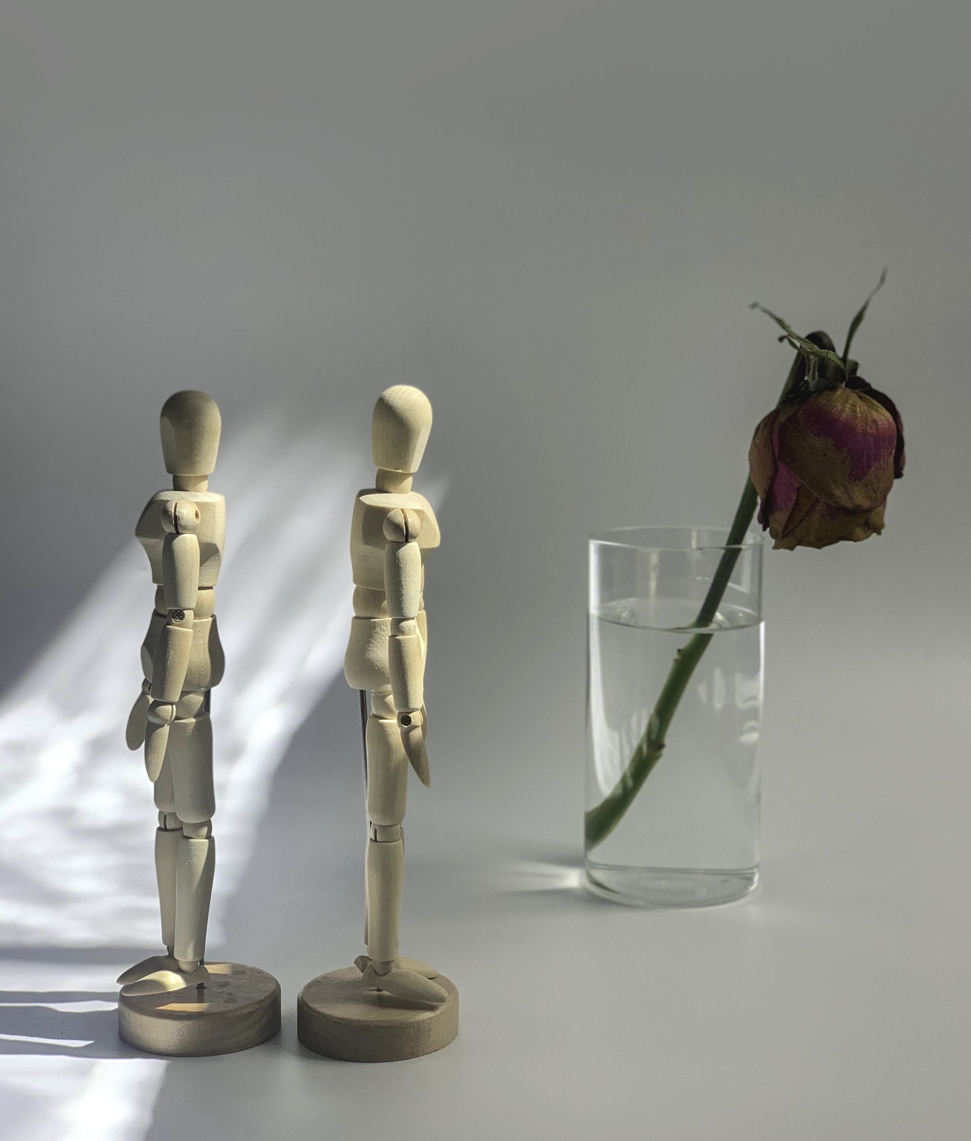 Two wooden mannequins are standing next to a glass of water and a dried rose.