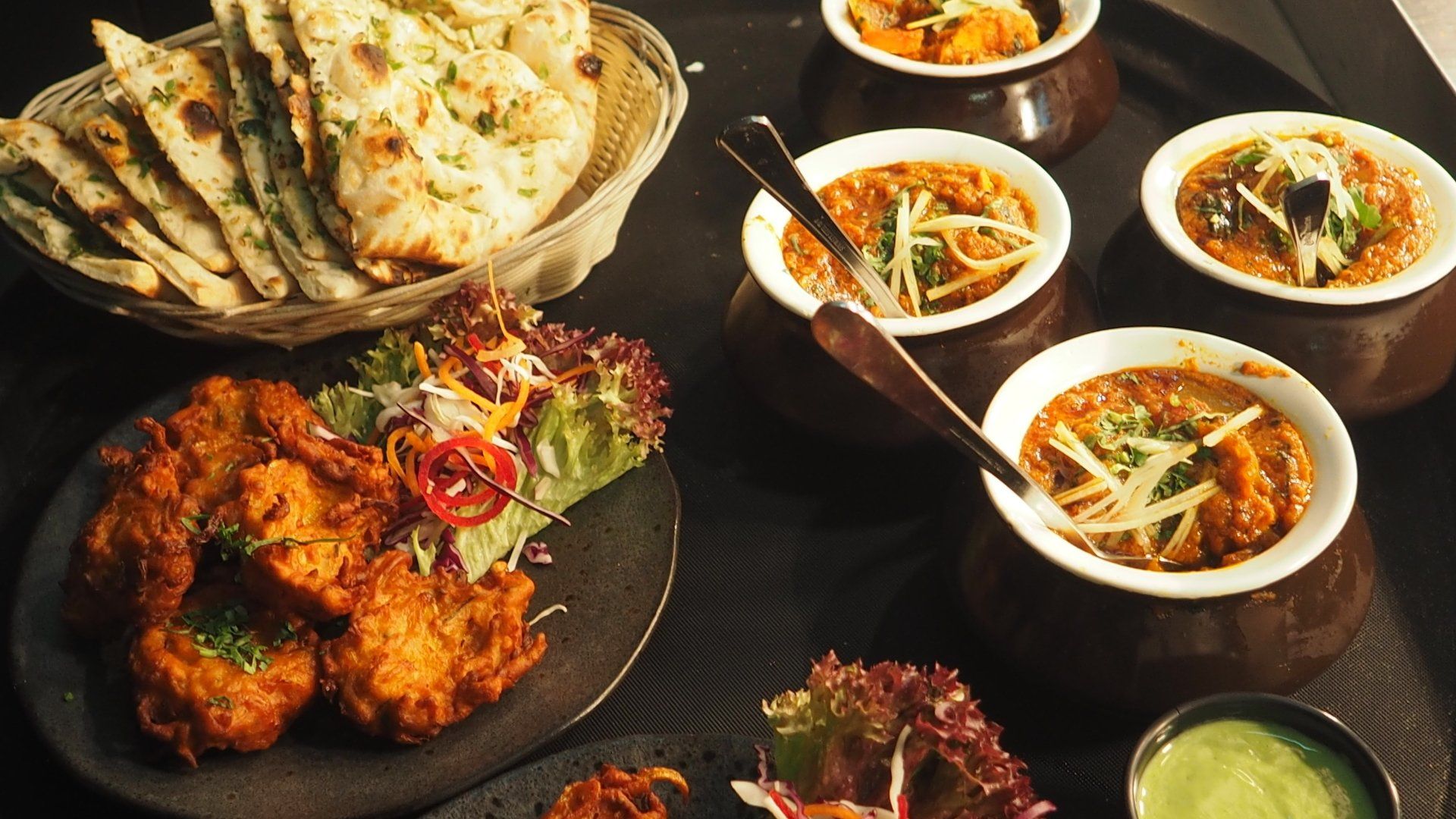 An image of several different types of food including curry, noodles, vegetables, chicken and naan bread. All of the food is either in white bowls and plates and are laid on a ceramic black worktop.