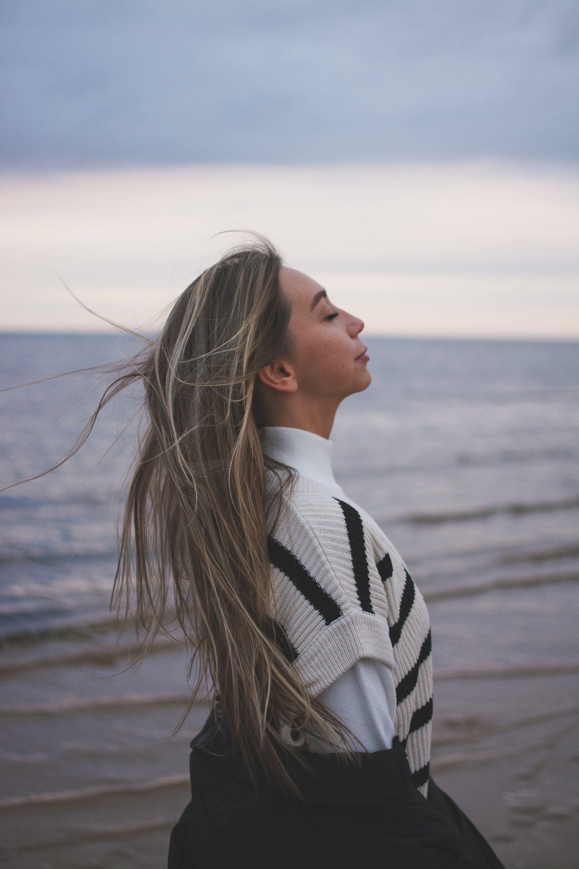 A woman is standing on a beach with her eyes closed and her hair blowing in the wind.