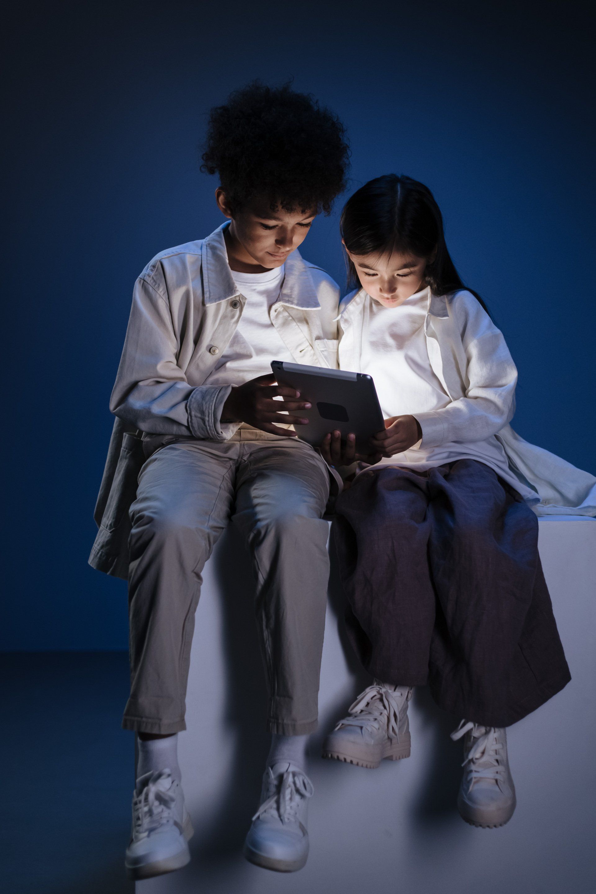 A boy and a girl are sitting on a cube looking at a tablet.