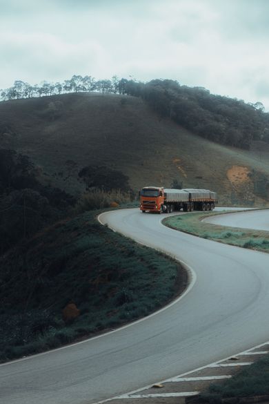 Orange semi-truck on winding asphalt road through green hills under a cloudy sky.