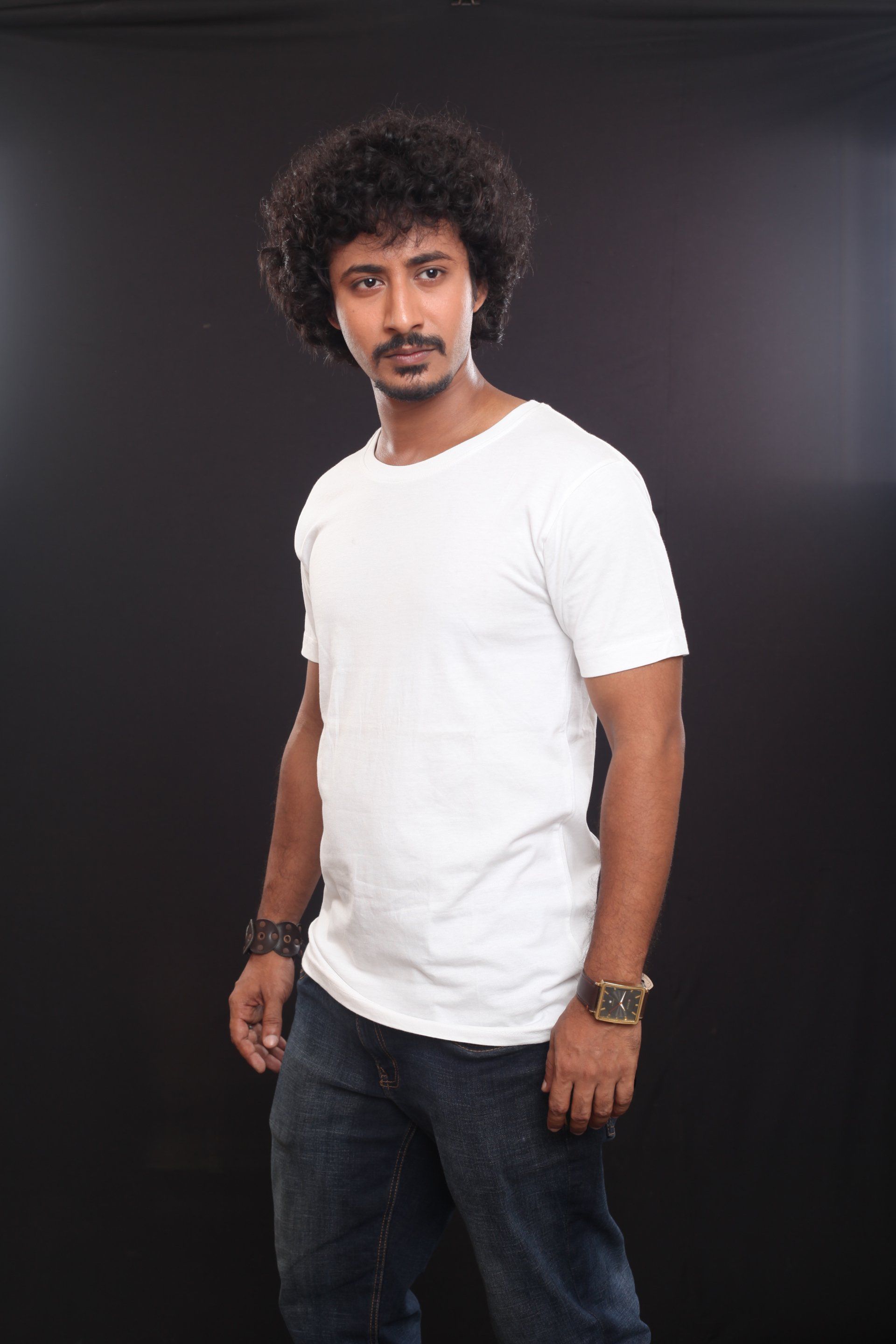 Man with curly hair in white t-shirt and jeans, looking to the side with a neutral expression, black backdrop.