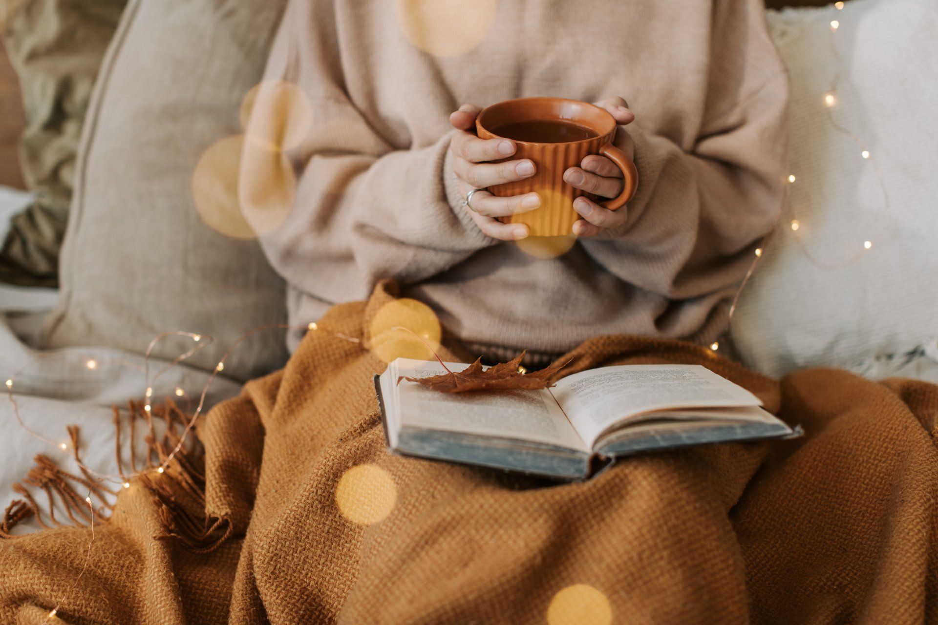 person holding a mug with a book in their lap cozying up on a couch with lights