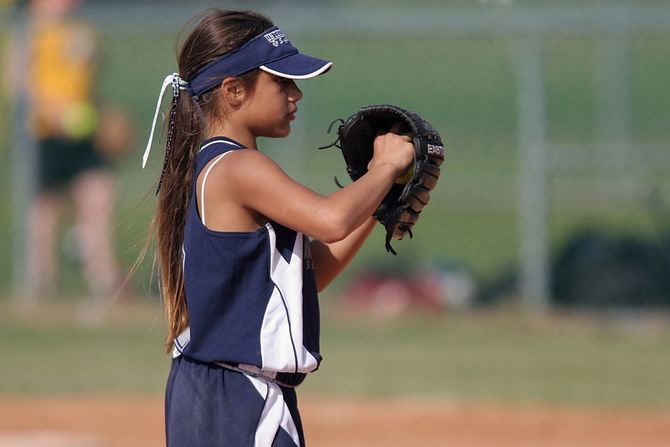 Young girl in softball uniform, wearing a visor, ready to pitch on a baseball field.