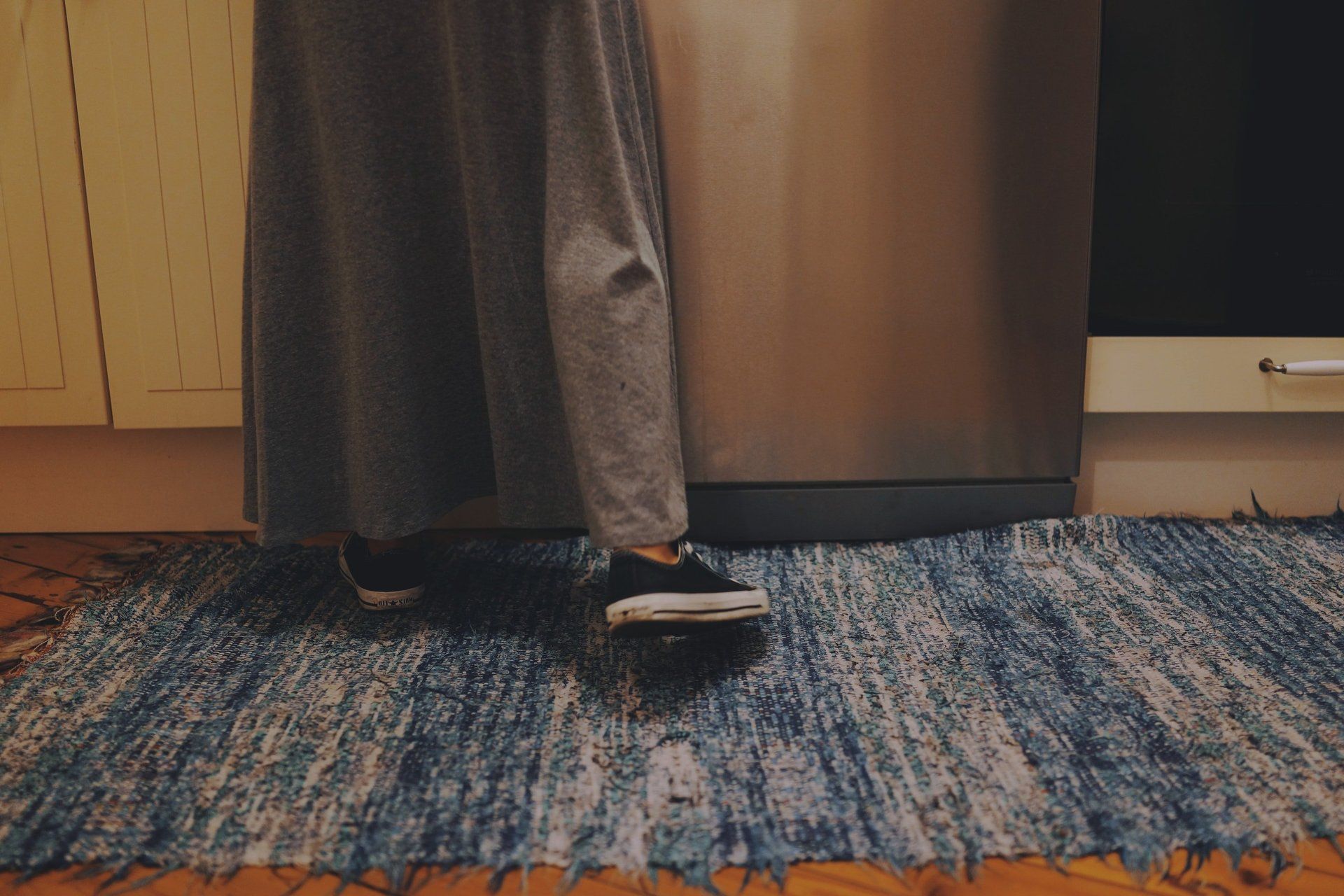 Person in a grey dress and black sneakers standing near a stainless steel refrigerator on a blue and white rug.