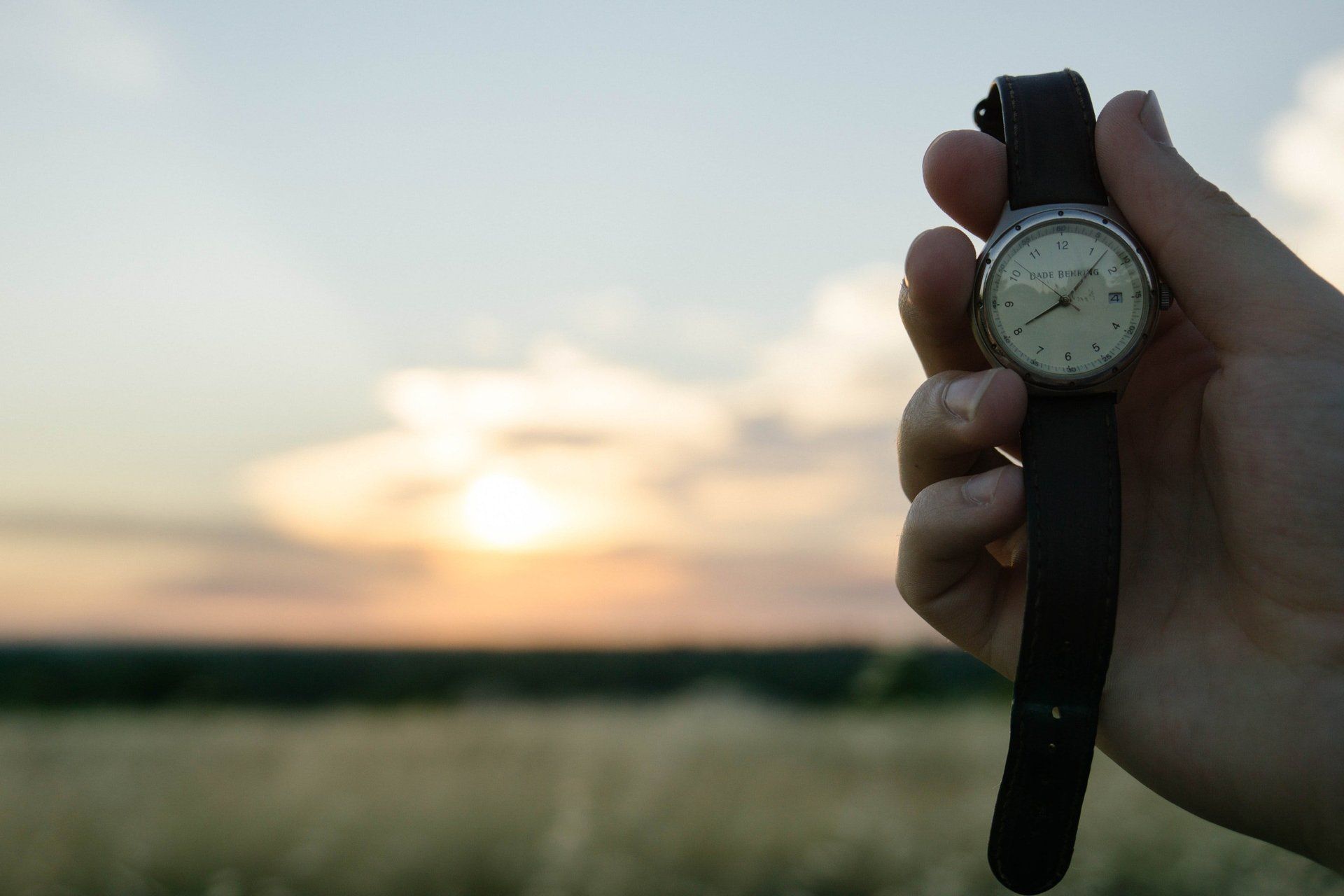 A person is holding a watch in front of a field at sunset.