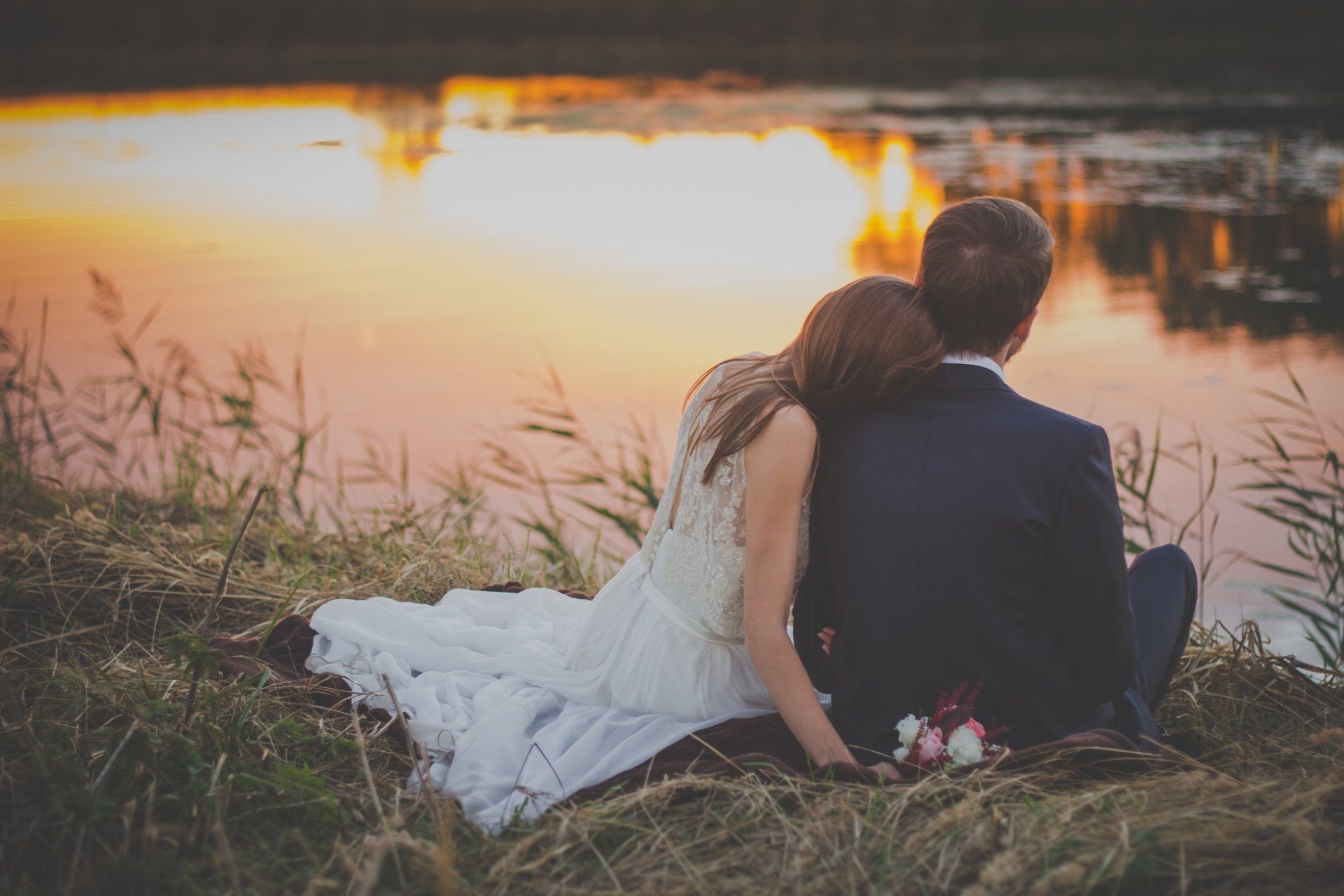 a bride and groom are sitting on the grass near a lake at sunset .