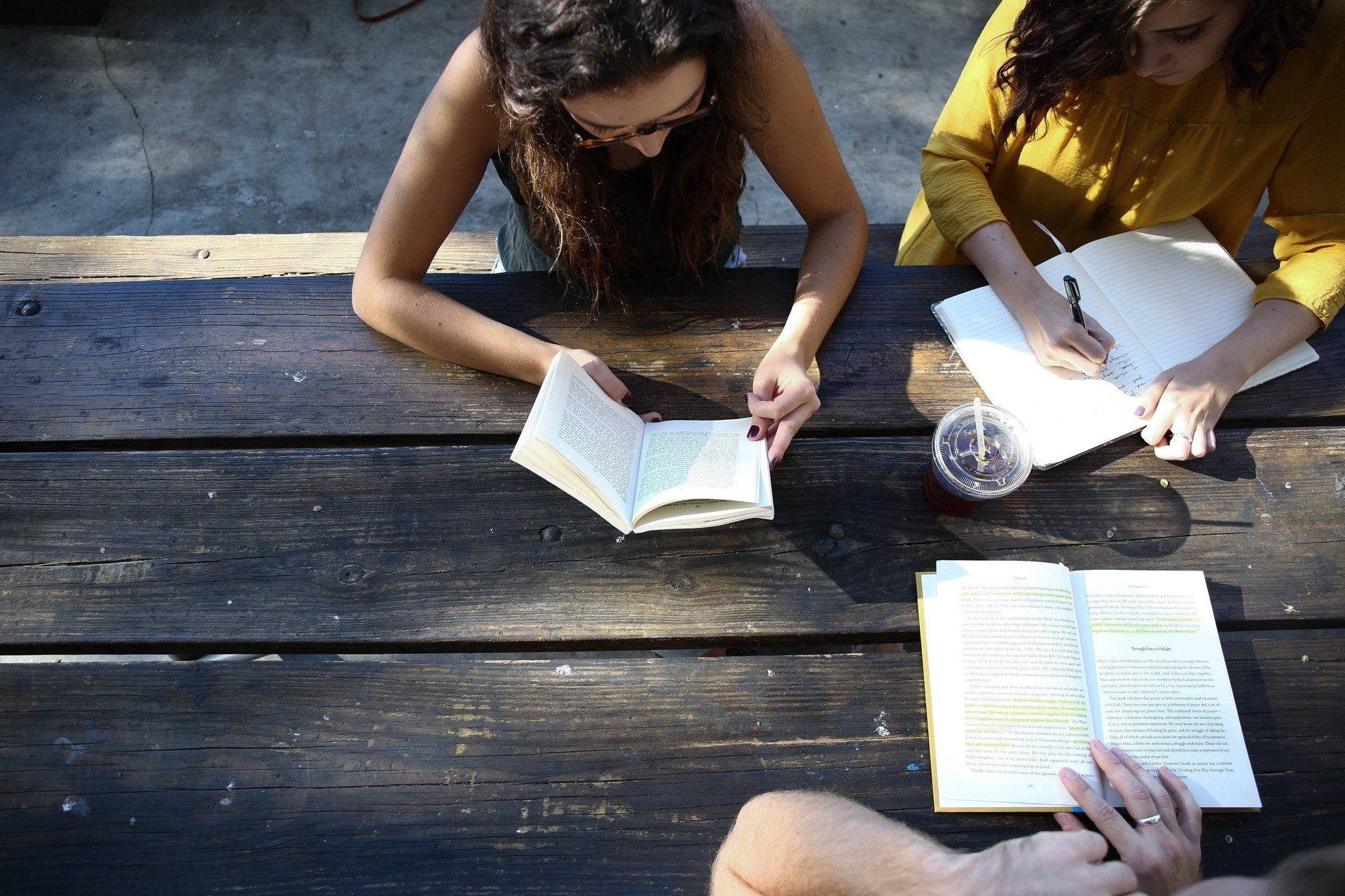 Three women are sitting at a picnic table reading books and writing in notebooks.