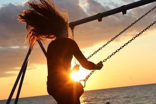 person with long hair swinging on swing set with ocean and sunset in background