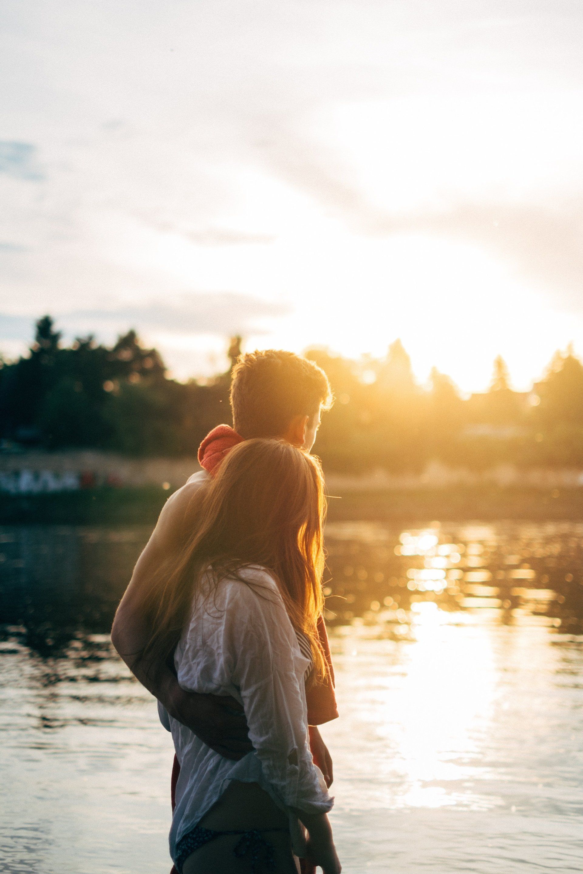 Young man and woman with arms around each other, walking beside and looking at a pond with sun setting in background.