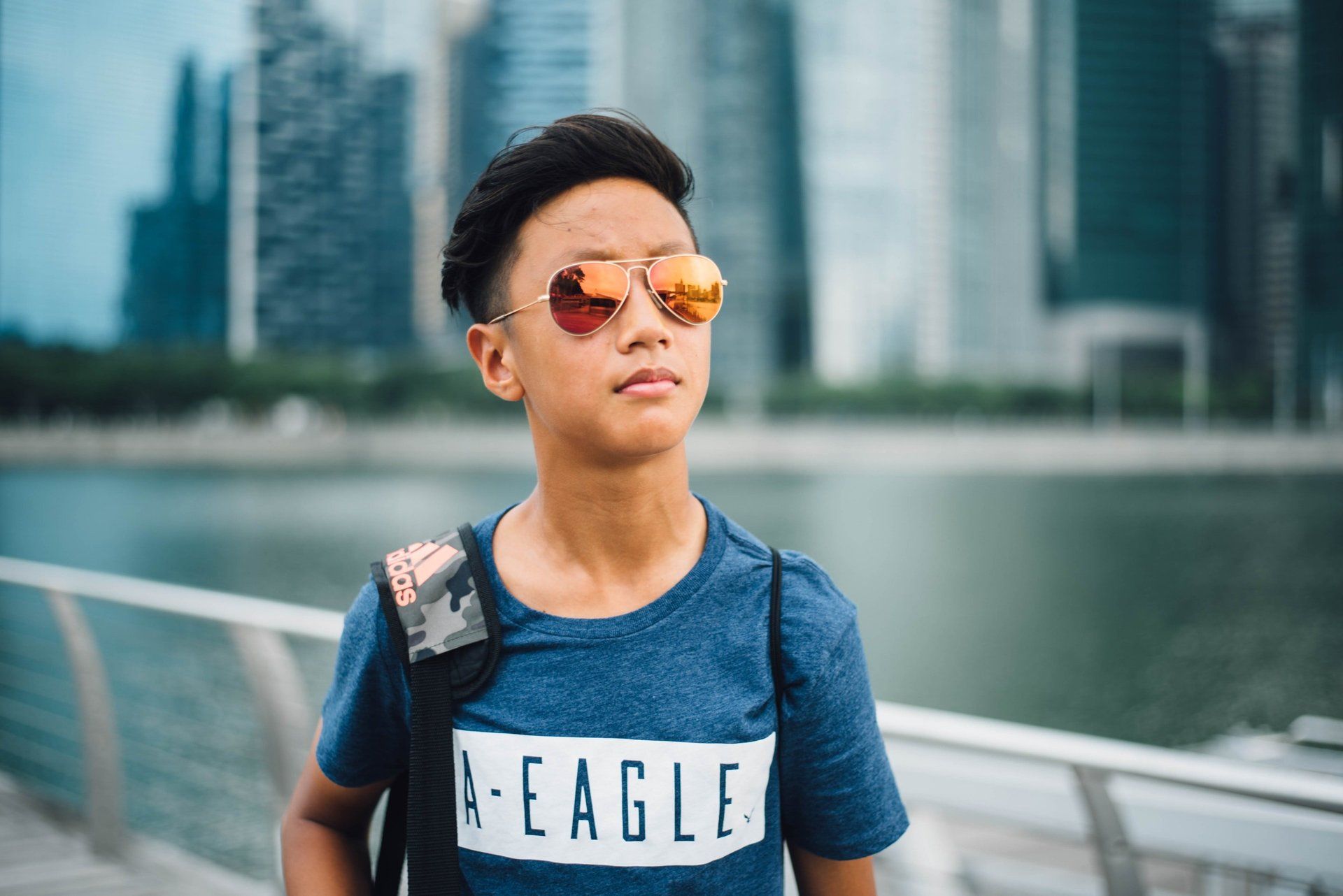 A young boy wearing sunglasses and a blue shirt is standing in front of a body of water.
